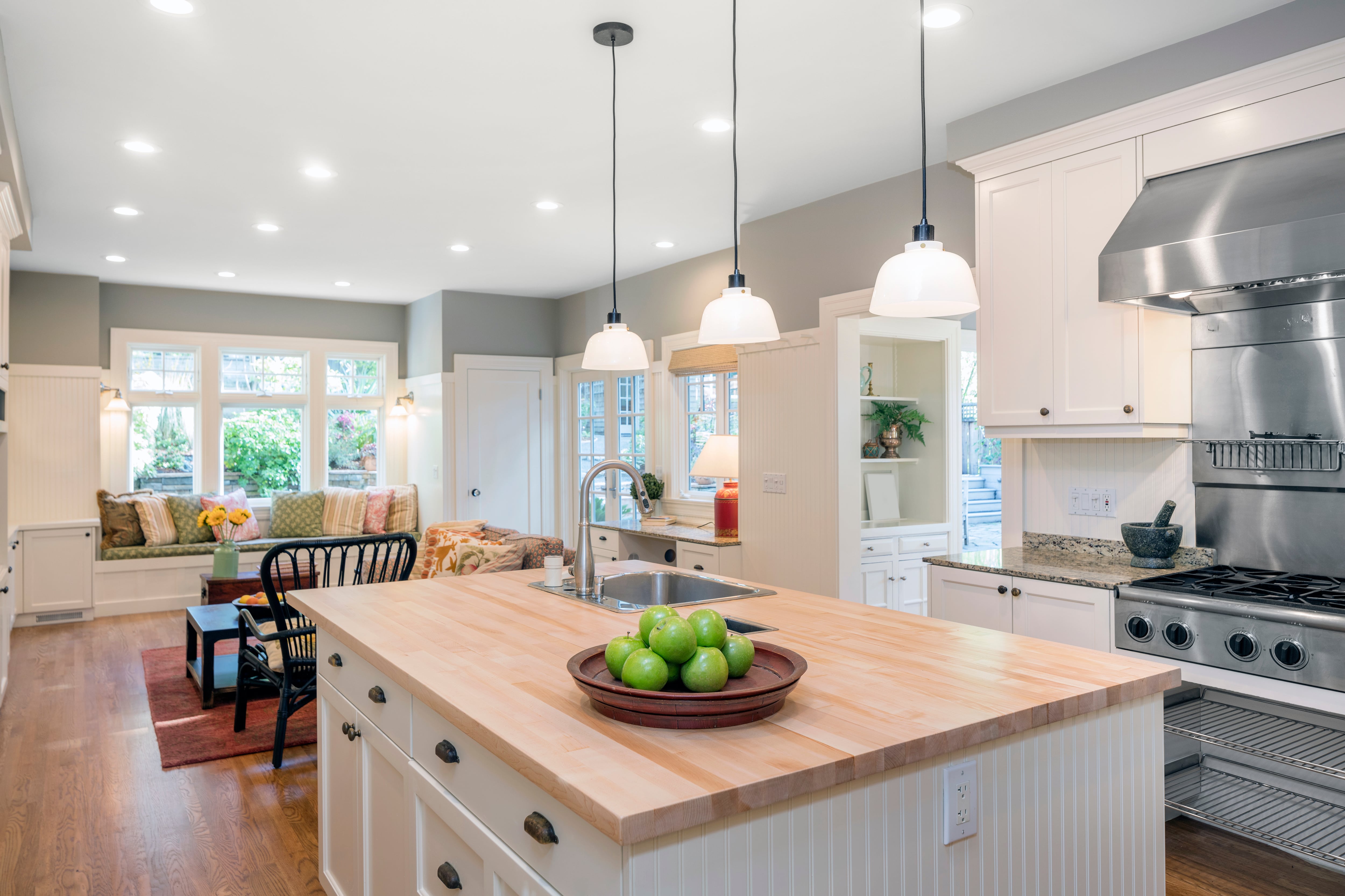 A bright, open-concept kitchen featuring a large wooden island with a bowl of green apples. The kitchen leads into a cozy living space with a built-in seating area by the windows, all illuminated by pendant lights.