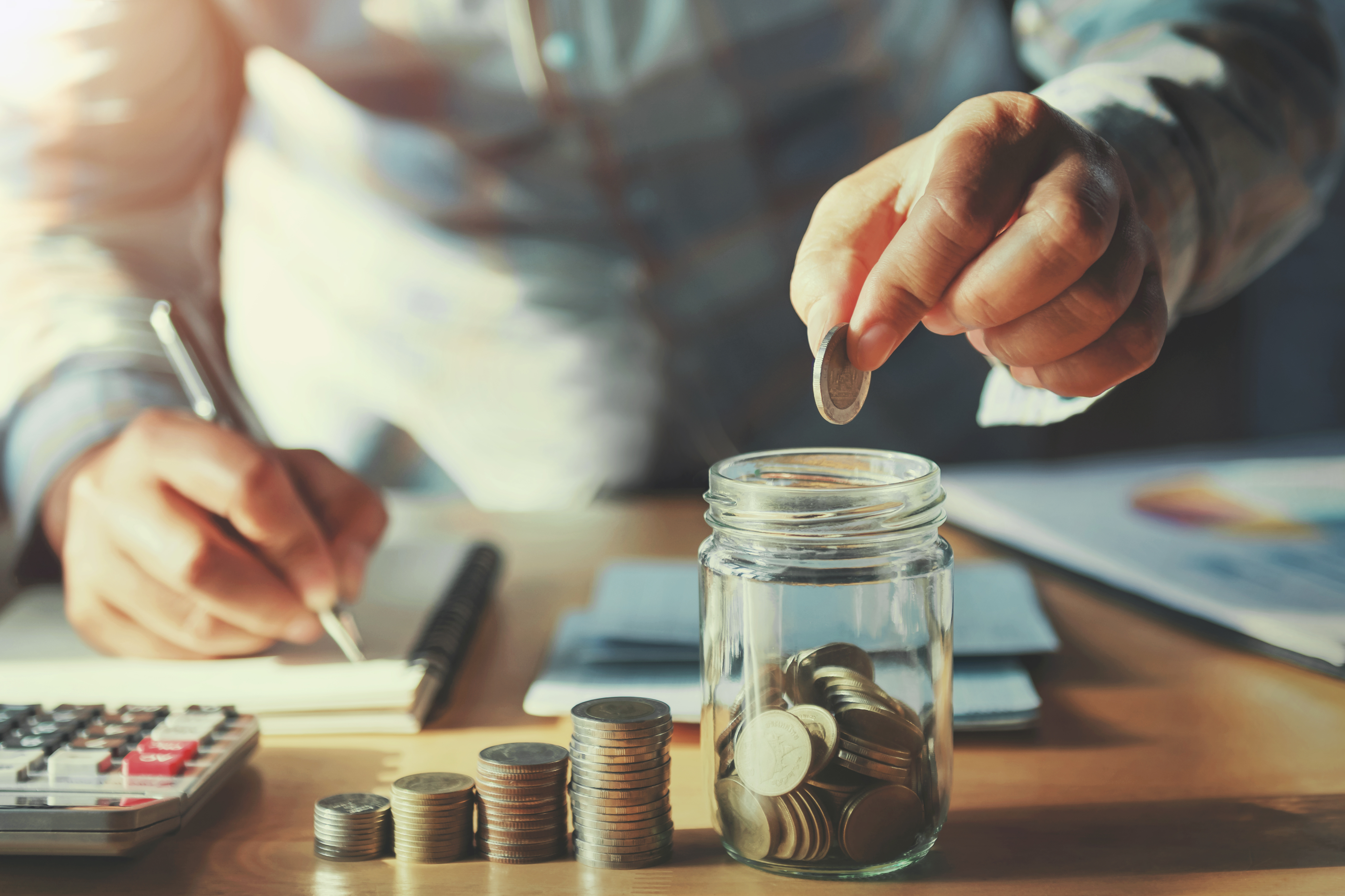 A person dropping coins into a glass jar, symbolizing saving money. A calculator and notes are visible on the table in the background.