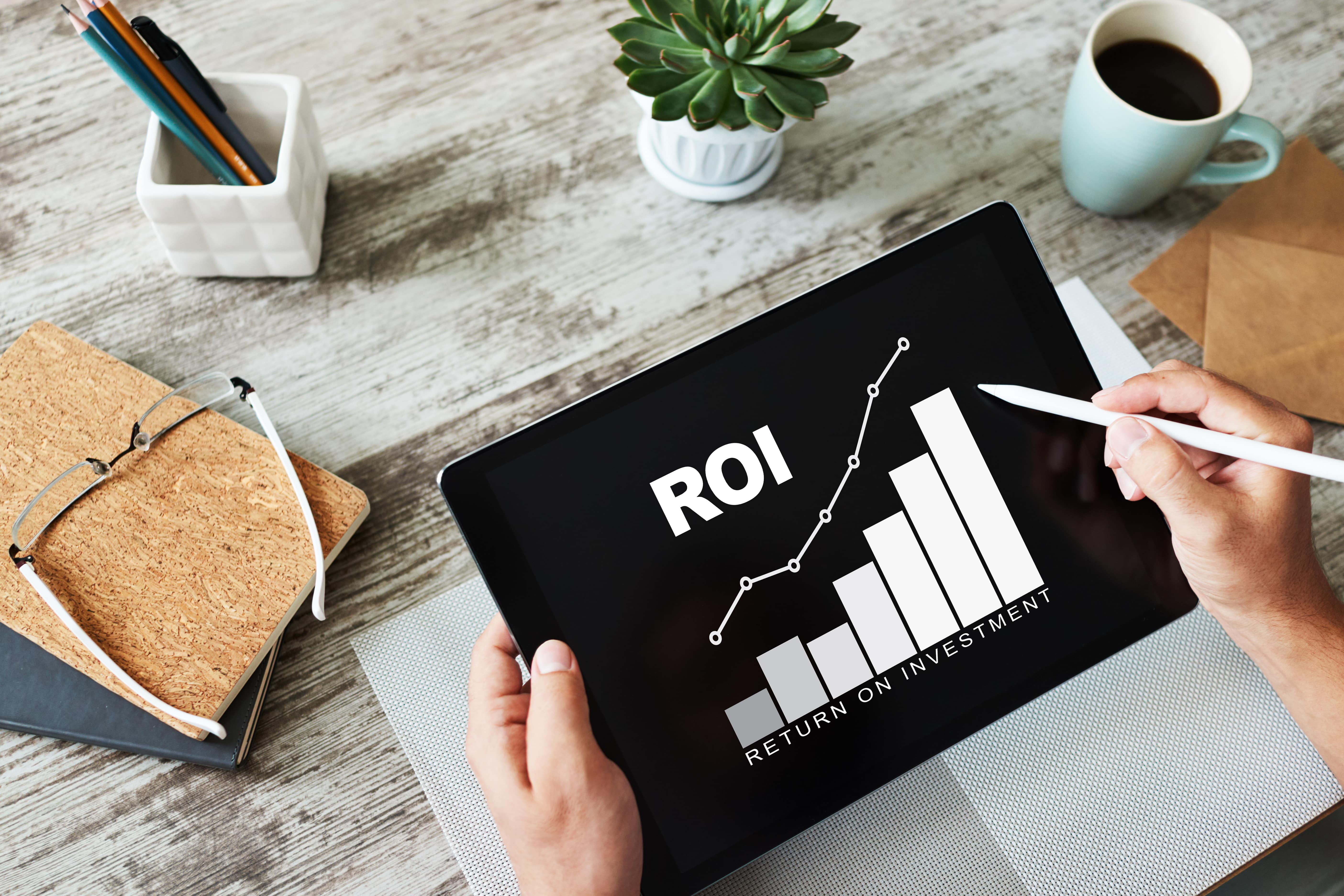 Hands holding tablet showing a bar chart labeled ROI (Return on Investment), surrounded by a work desk setup.