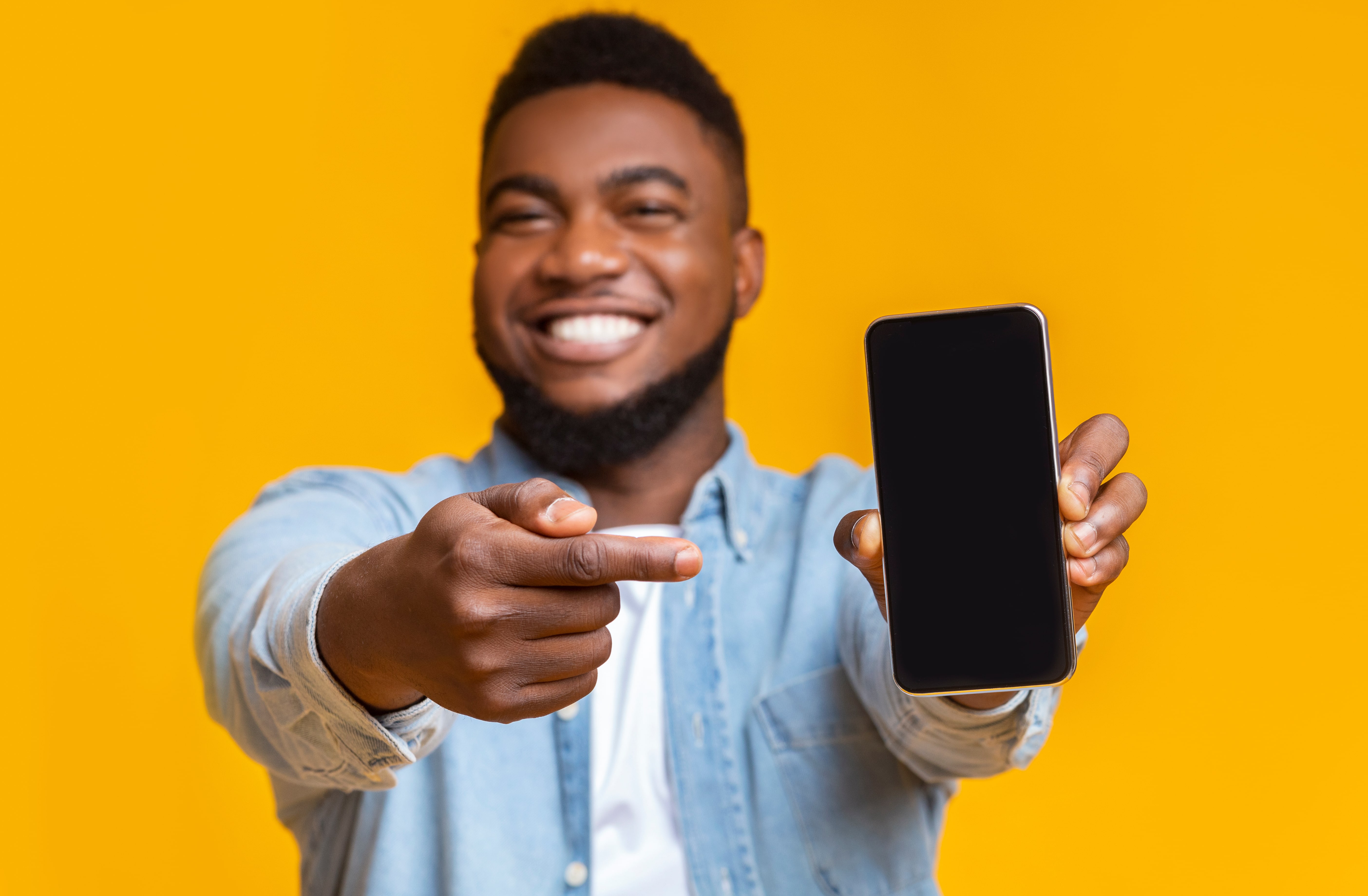 Smiling man holding and pointing at a smartphone with a blank screen, against a bright yellow background.