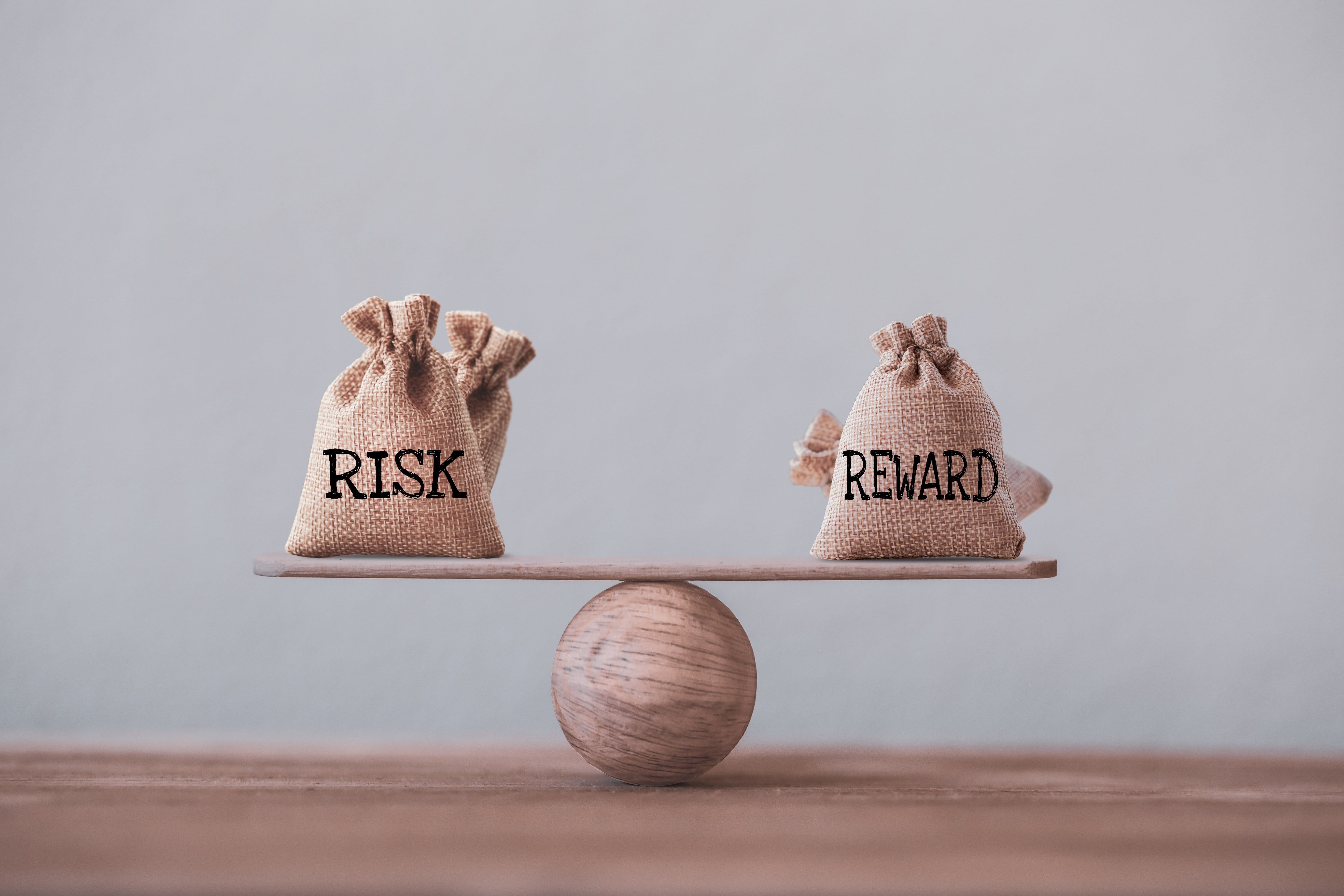 Two small burlap bags labeled ‘Risk’ and ‘Reward’ balanced on a wooden seesaw, illustrating the balance investors must consider with the BRRRR method.