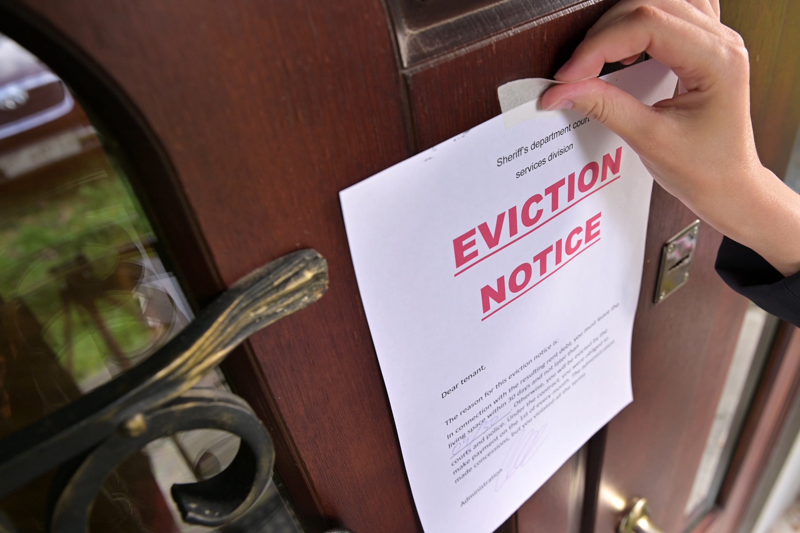 A close-up of a person’s hand taping an eviction notice to a wooden door, illustrating the formal posting process.