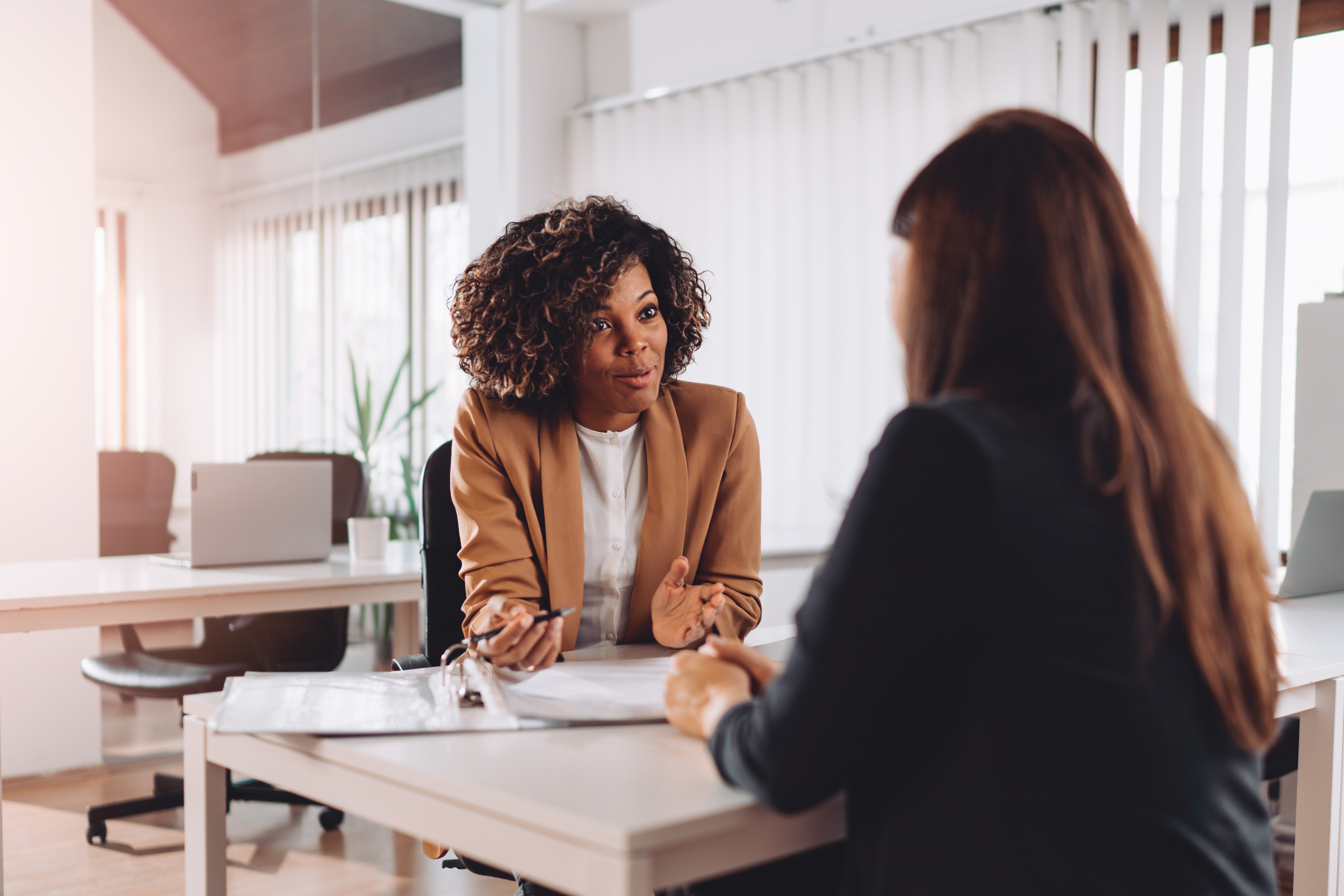 A businesswoman with curly hair and a tan blazer is sitting at a desk, talking with another person in a modern, well-lit office.