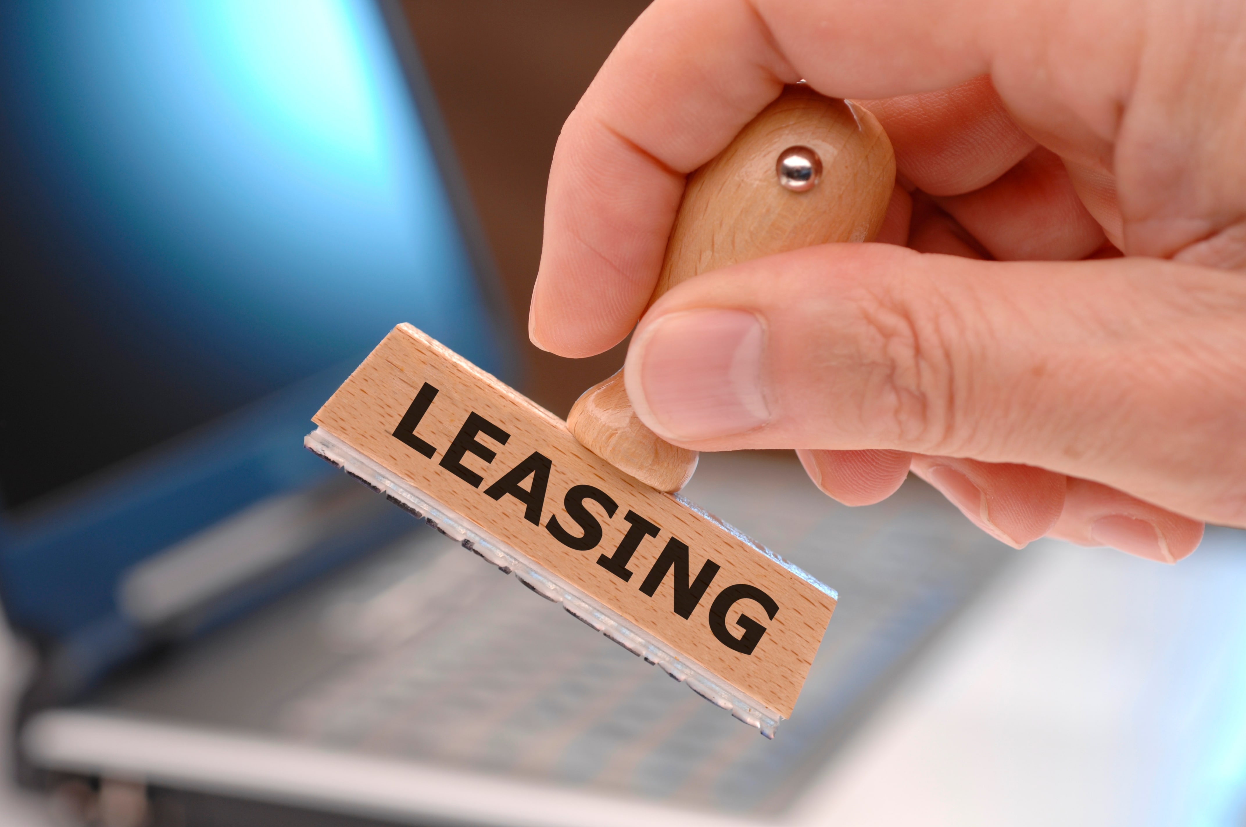 Hand holding a wooden stamp labeled “LEASING” in bold letters, with a blurred laptop in the background.
