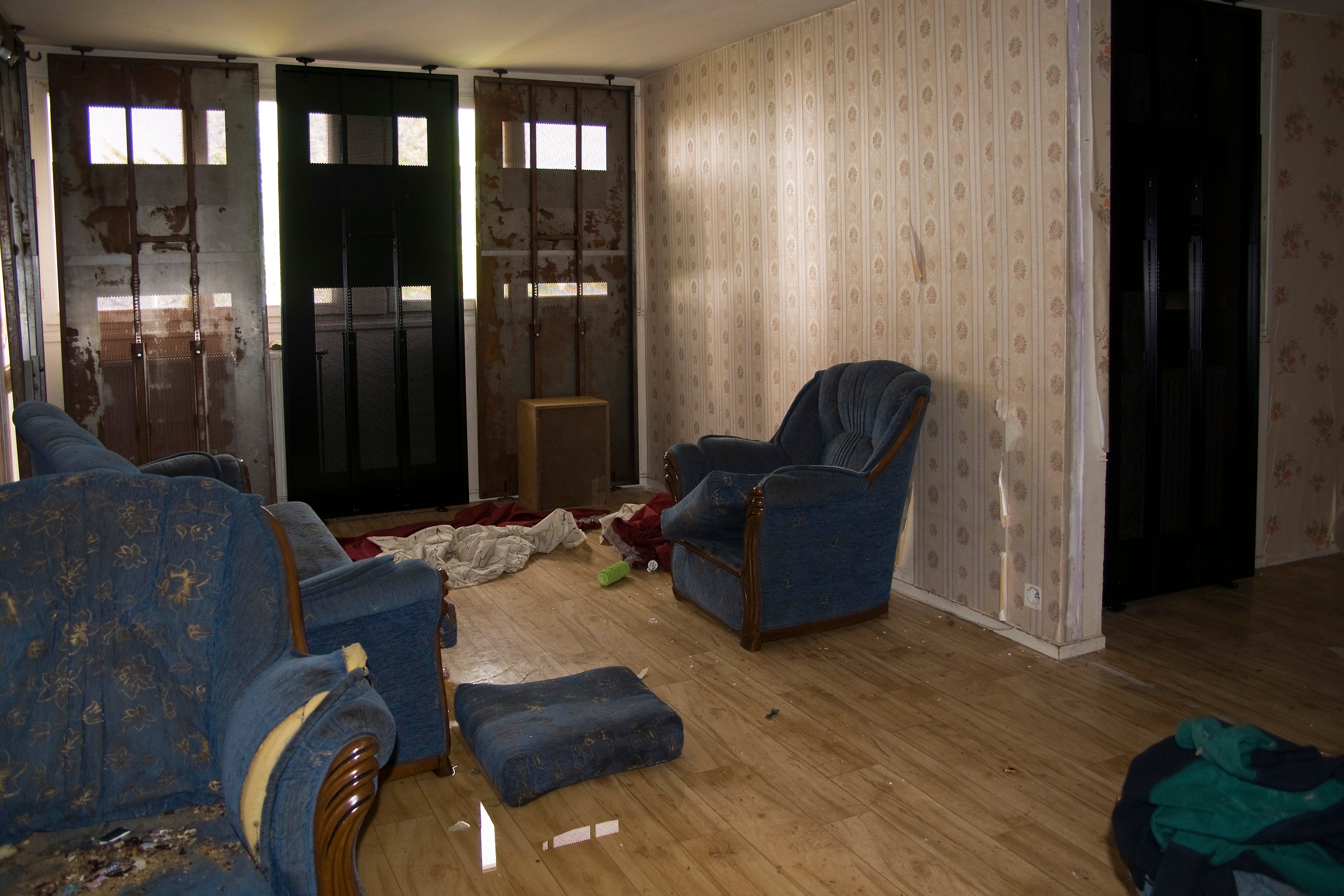 A neglected living room with tattered furniture, scattered debris, and boarded-up windows, showing signs of abandonment and potential squatter activity.