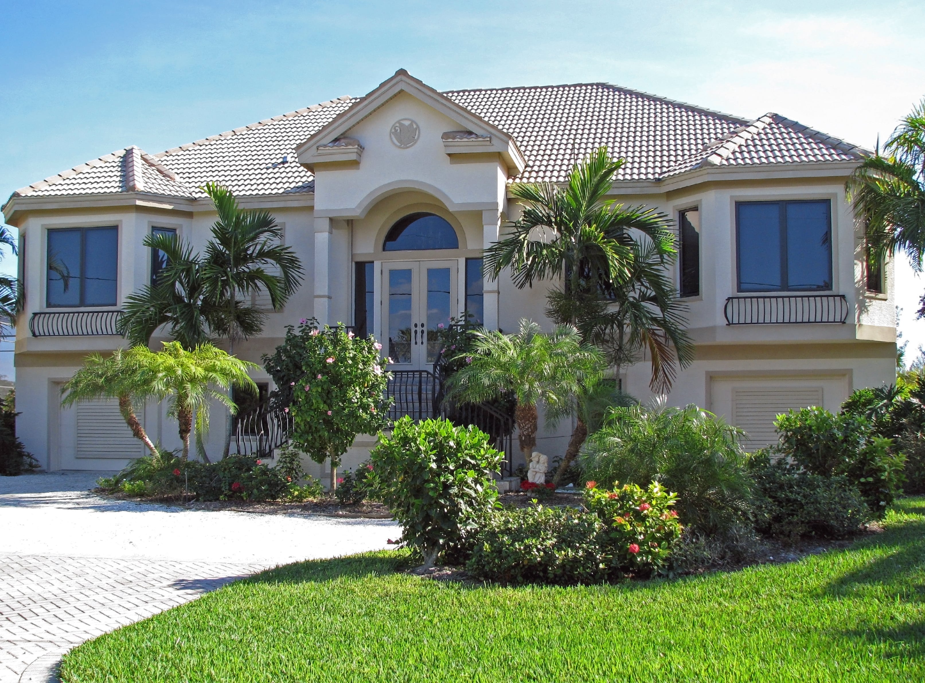 A luxurious two-story Mediterranean-style home with a beige stucco exterior, arched entryway, and a well-manicured lawn with tropical landscaping, including palm trees and flowering bushes, under a bright blue sky.
