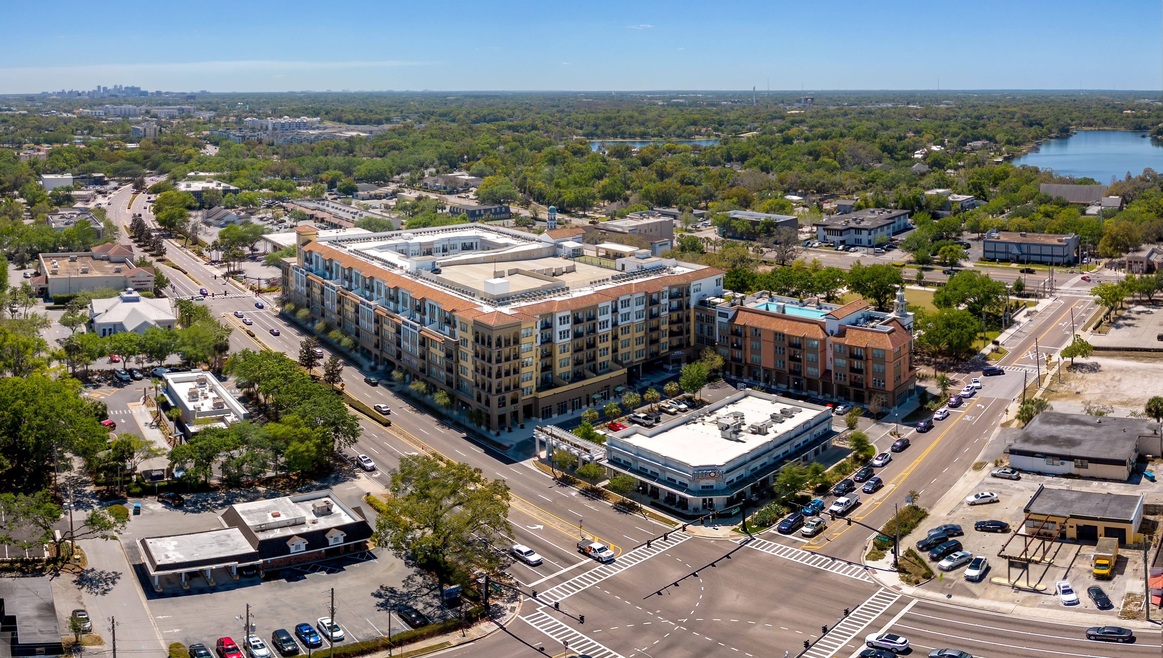 A high-angle view of a modern mixed-use building in Winter Park, Florida, showcasing a vibrant urban neighborhood with tree-lined streets, businesses, and residential buildings. Downtown Orlando is visible in the distance.