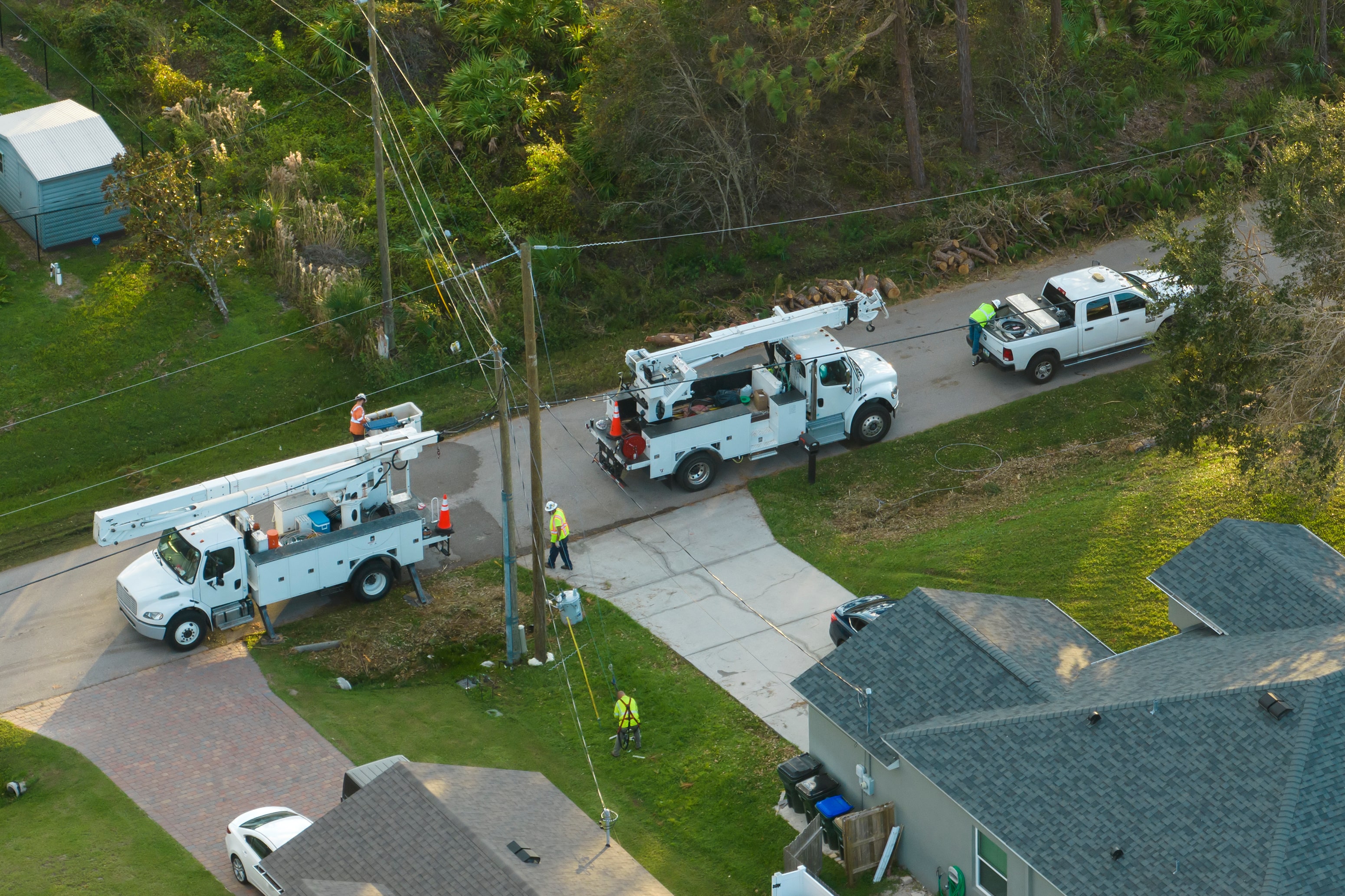 aerial view of utility workers and their trucks parked on a residential street. Two white utility trucks are stationed near a power line, with workers actively working to restore service. A third vehicle, a pickup truck, is parked nearby, and one worker can be seen walking in a safety vest. This scene suggests maintenance or repairs following a storm, likely focusing on restoring power to the area. The surrounding area includes trees, grass, and residential homes.