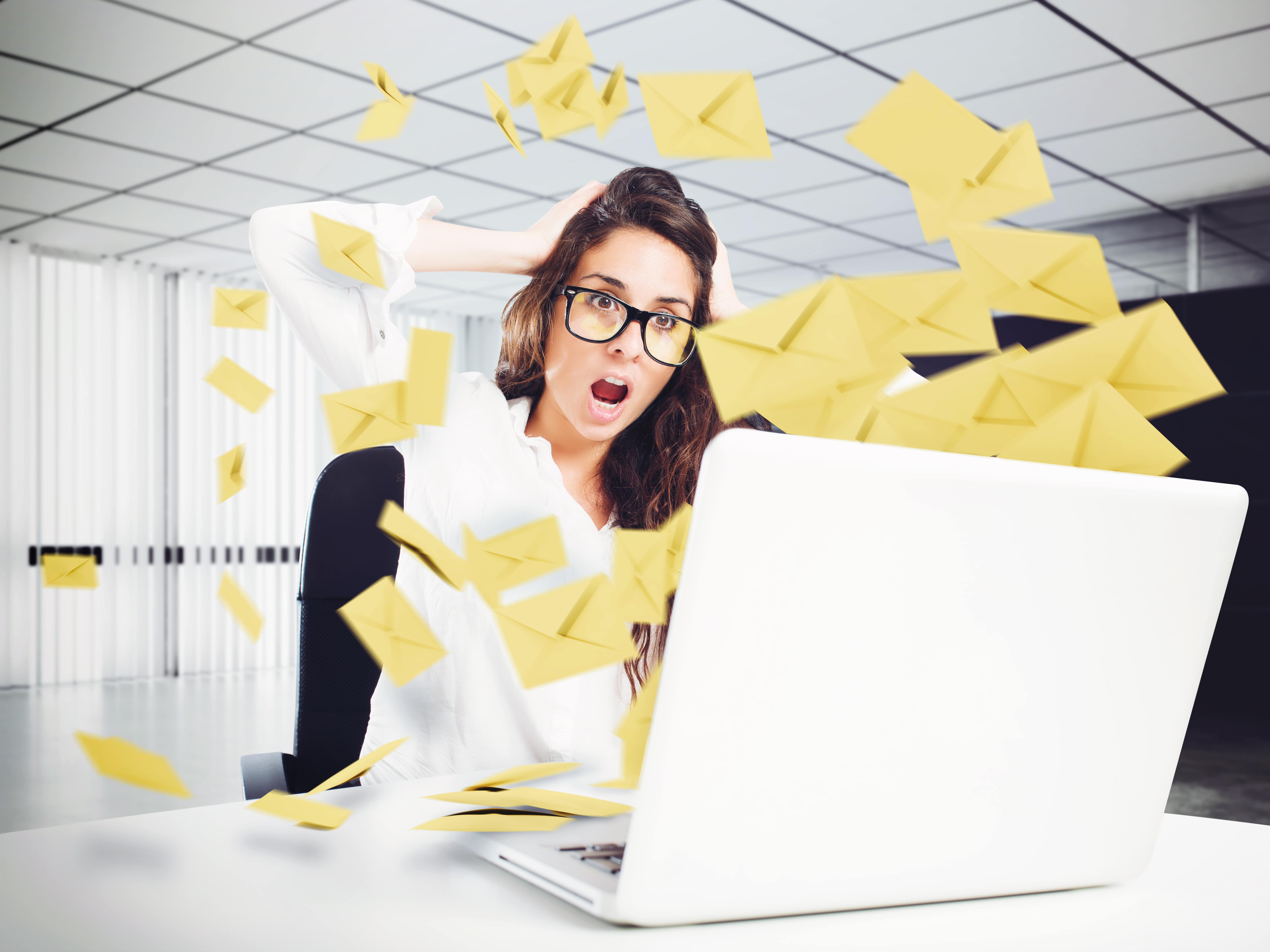 Stressed woman wearing glasses, sitting at a desk with a laptop, as yellow email envelopes appear to fly toward her, representing email overload.