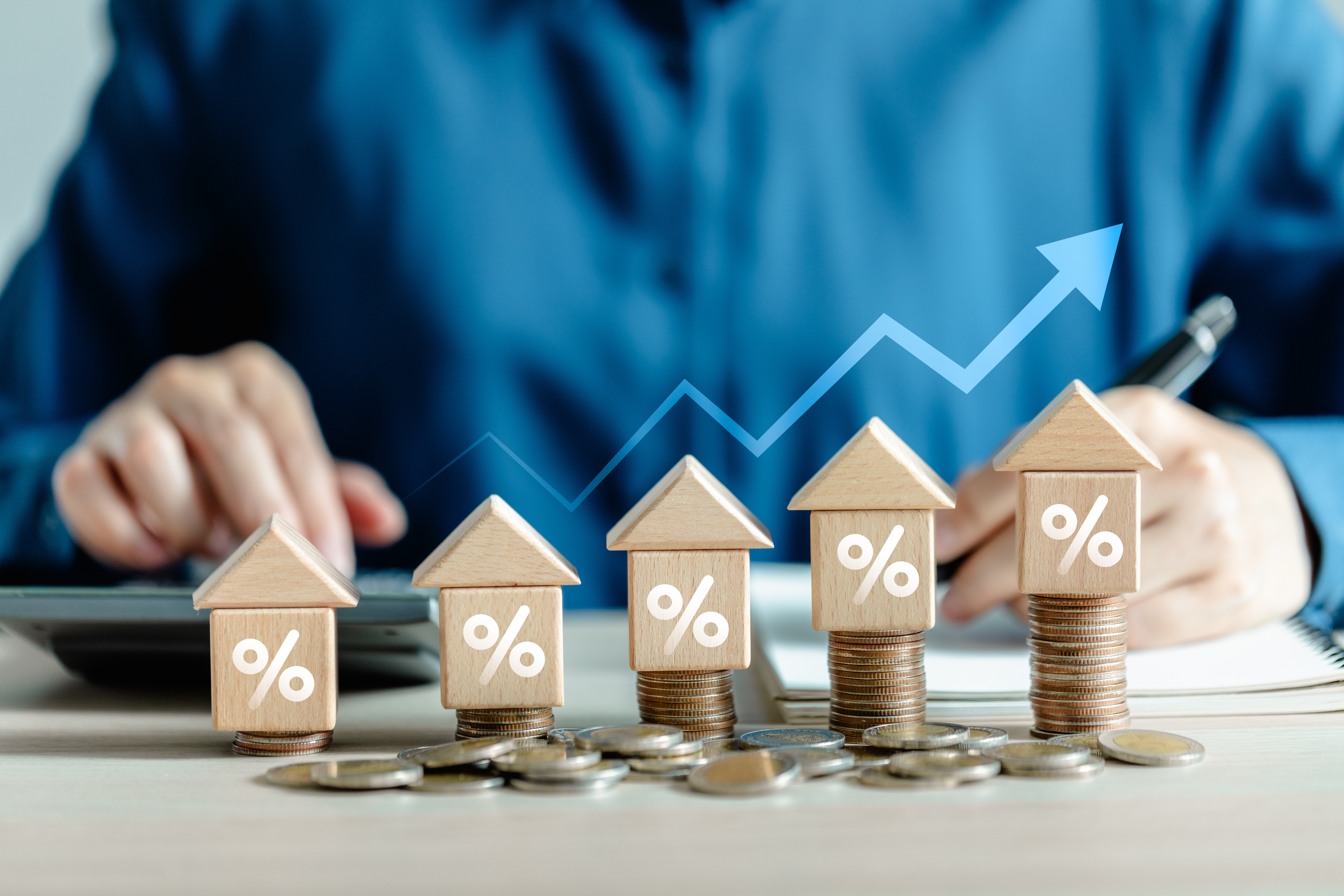 Wooden house models with percentage symbols stacked on coins, representing real estate investment growth. A graph with an upward trend is in the background.