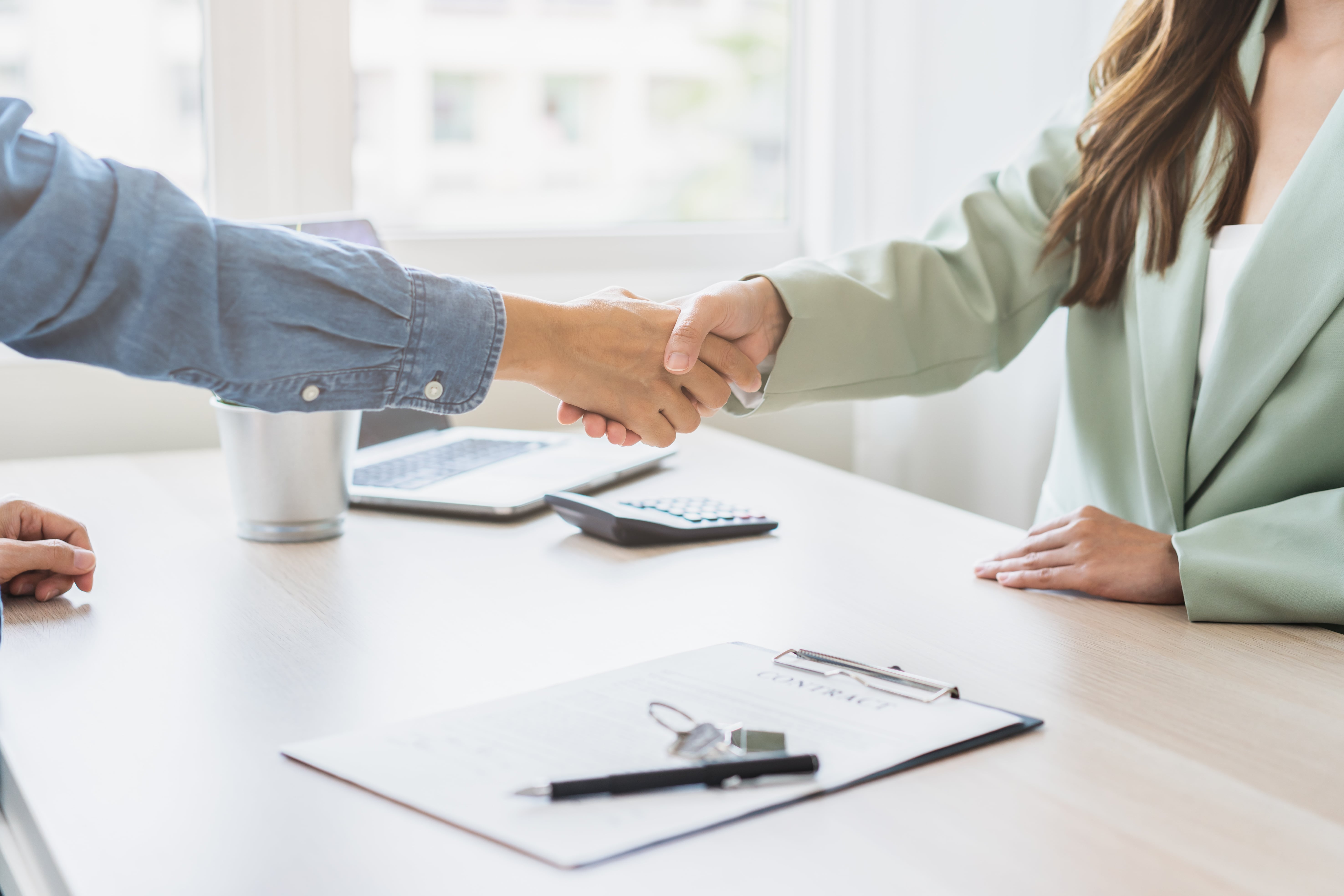 Two people shaking hands over a signed contract with house keys and a pen on the table, symbolizing a successful lease agreement.