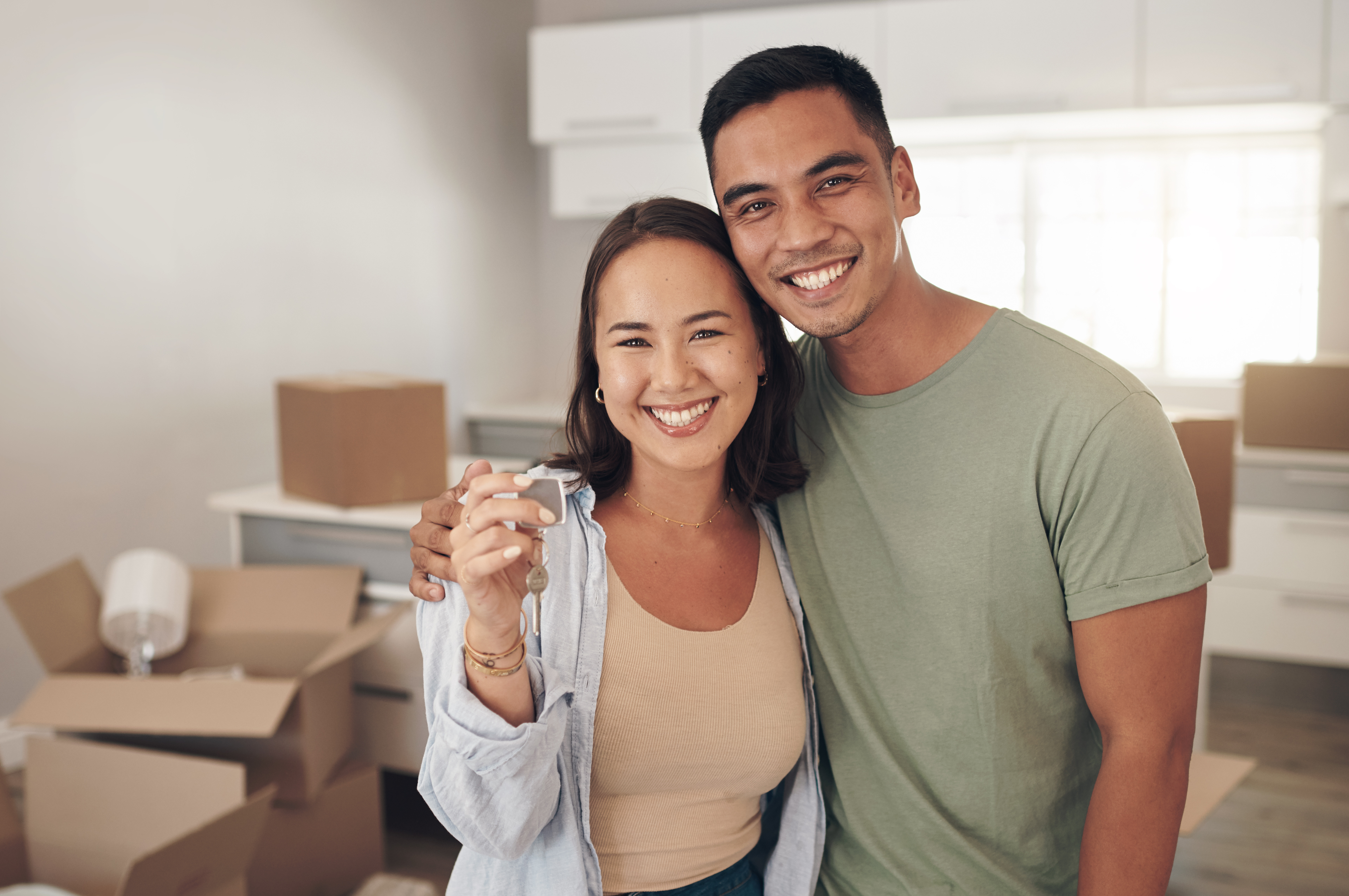 A happy couple stands in their new home, smiling at the camera. The woman is holding a set of house keys, symbolizing the moment they have moved in. Behind them are packed moving boxes, with the kitchen visible in the background, suggesting they are in the process of settling into their new home.