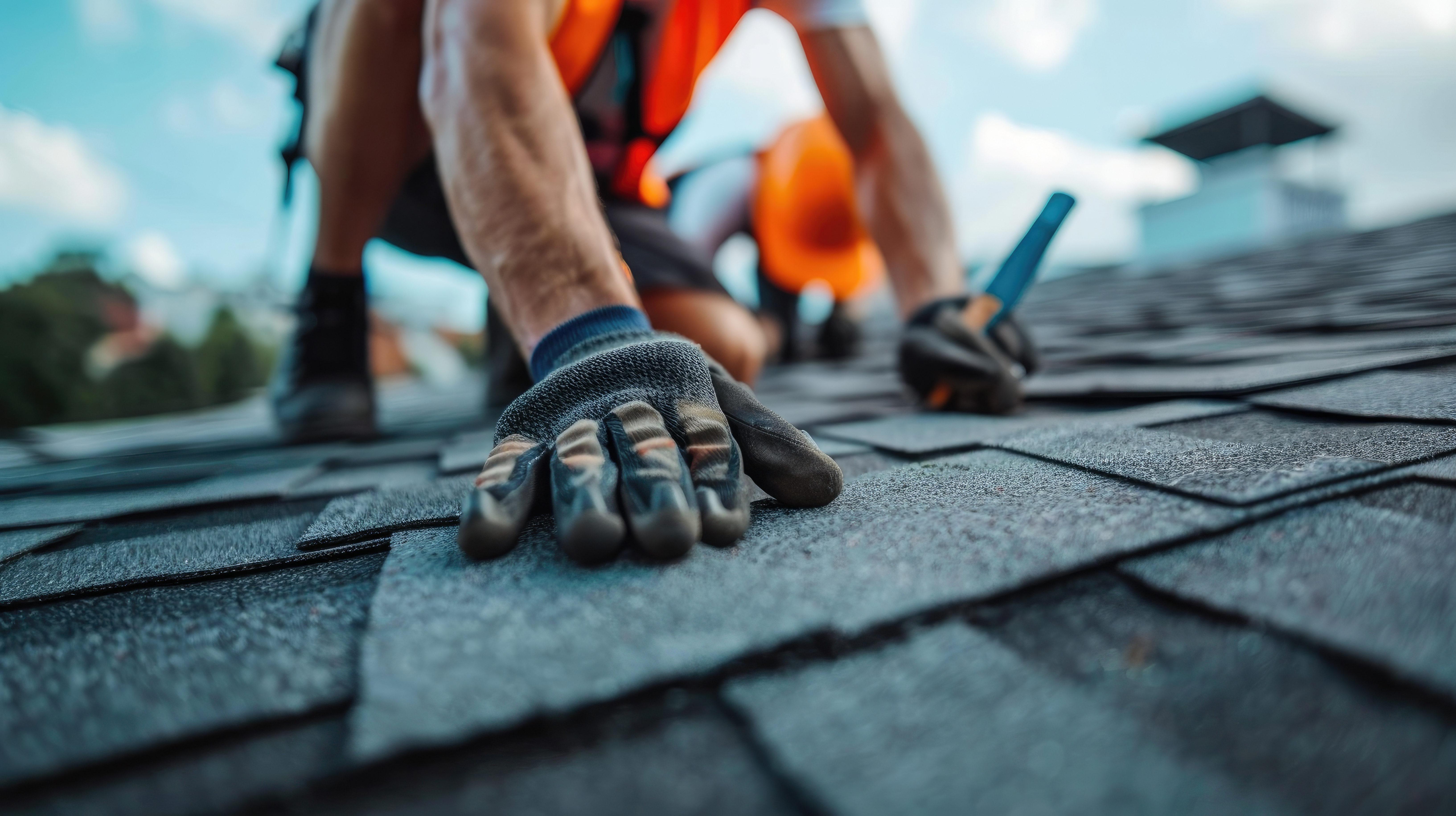 A close-up of a gloved hand carefully placing shingles on a roof, with another worker visible in the background. Both workers wear safety vests.