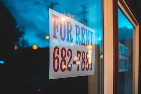 A 'FOR RENT' sign in a window with a blurred evening background.