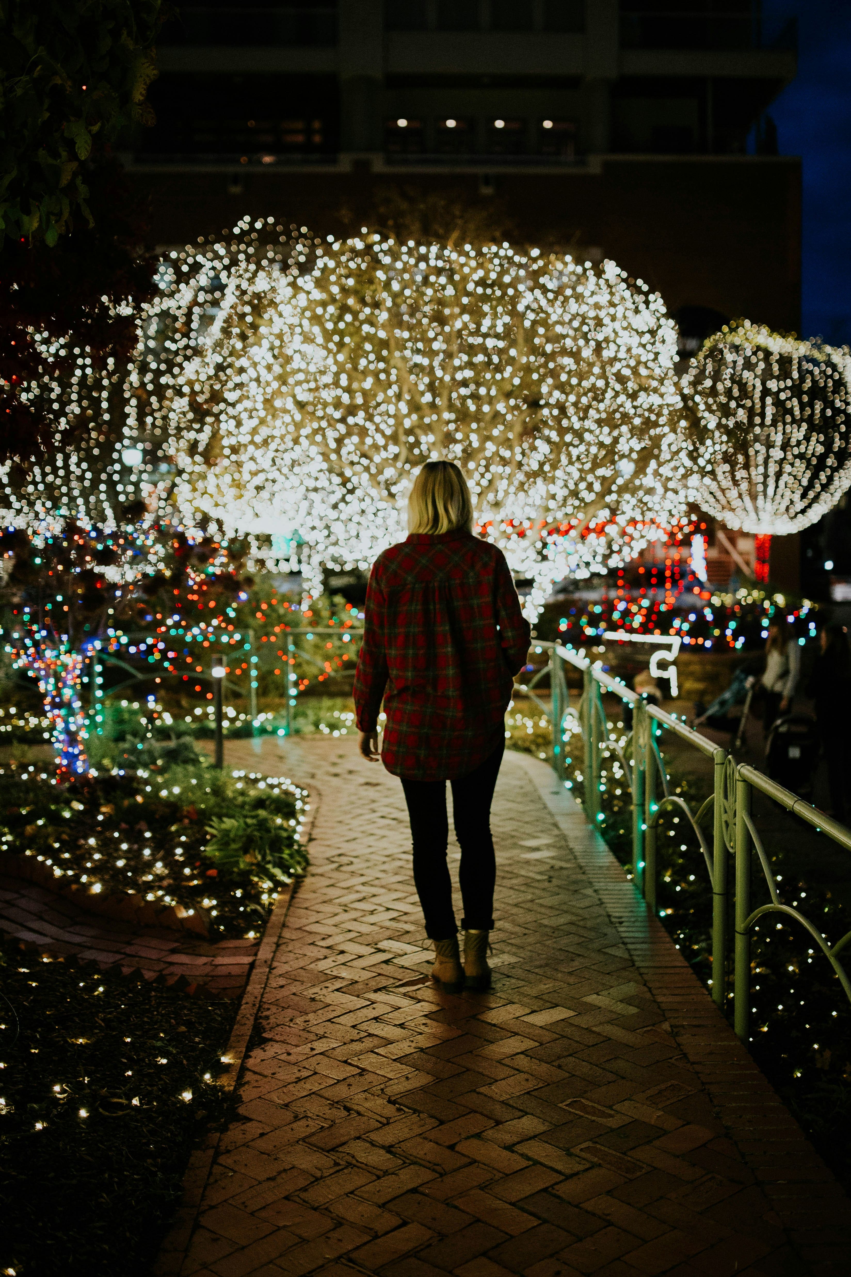 A woman wearing a plaid red shirt walking along a beautifully lit pathway adorned with twinkling holiday lights on trees and bushes. Photo by Brooke Cagle on Unsplash