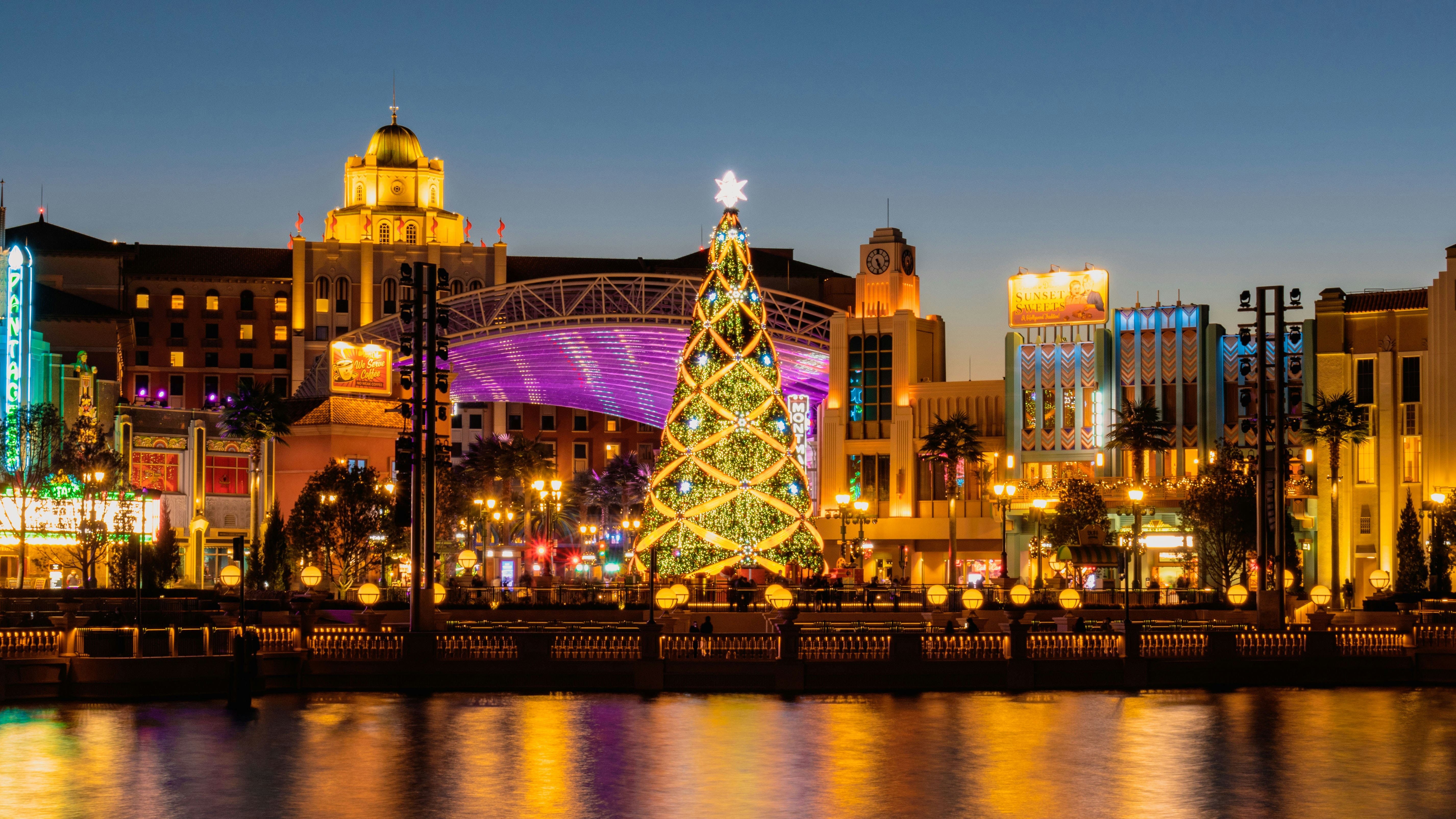 A large illuminated Christmas tree at a vibrant city square, surrounded by colorful lights and reflecting on the water at dusk. Photo by Mingfang on Unsplash 