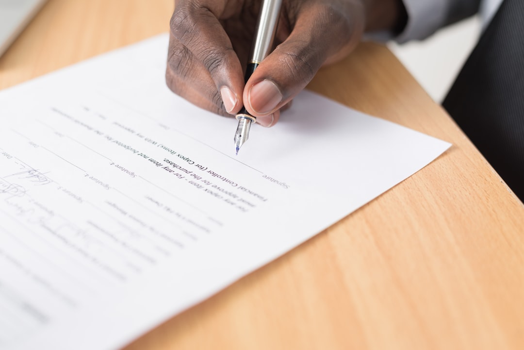 A close-up of a person signing a document with a silver pen on a wooden desk.