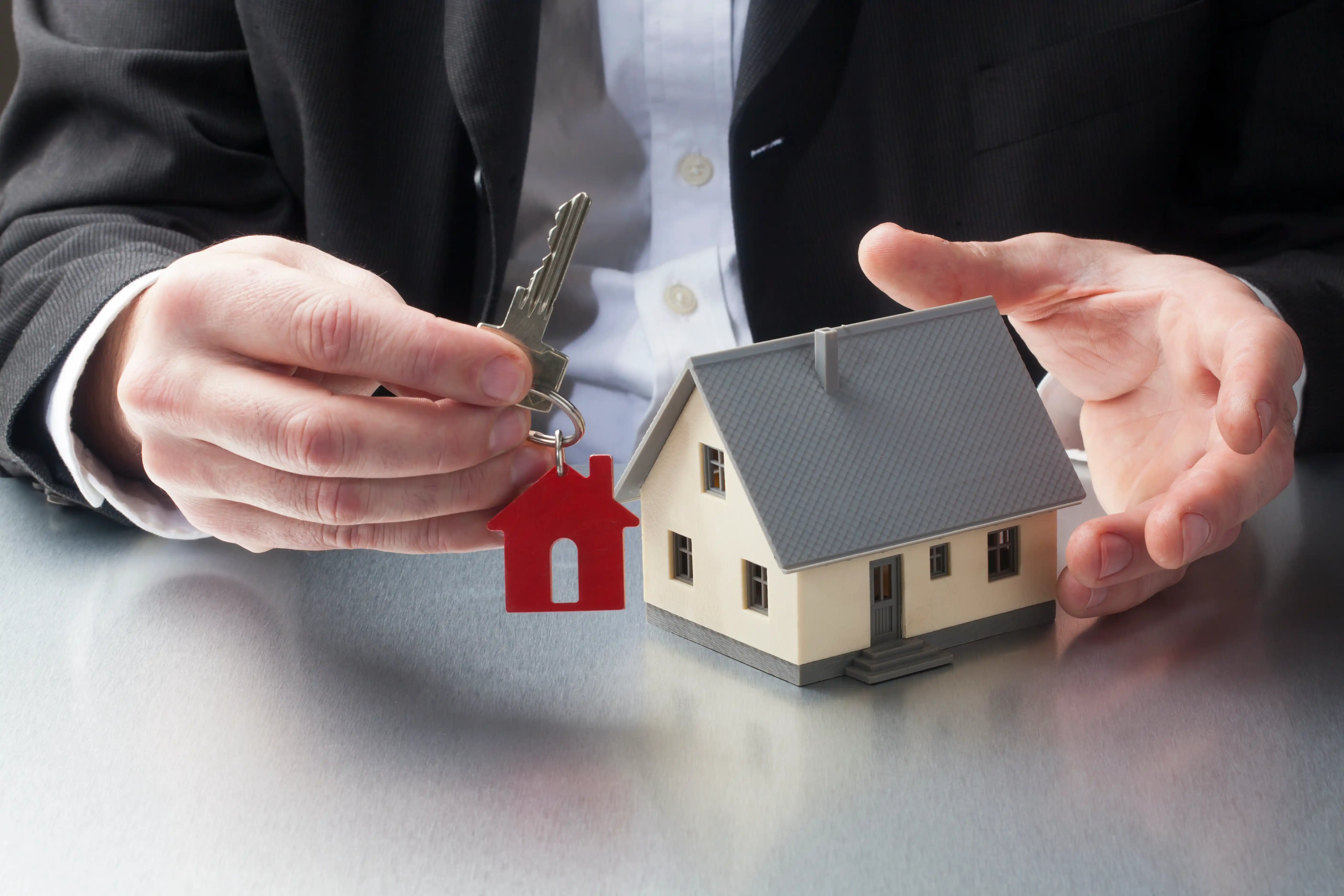 Businessperson handing over house keys next to a model home, representing real estate investment and property management transactions.